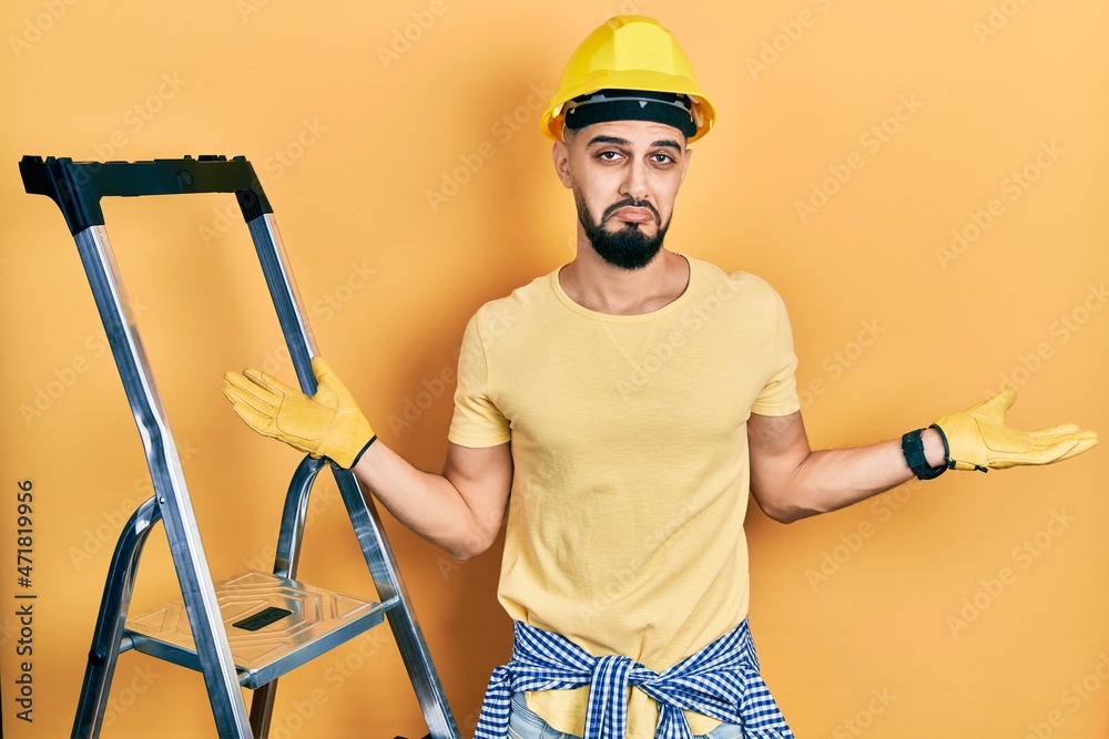 Handsome man with beard by construction stairs wearing hardhat smiling showing both hands open palms, presenting and advertising comparison and balance Handsome man with beard by construction stairs wearing hardhat smiling showing both hands open palms, presenting and advertising comparison and balance