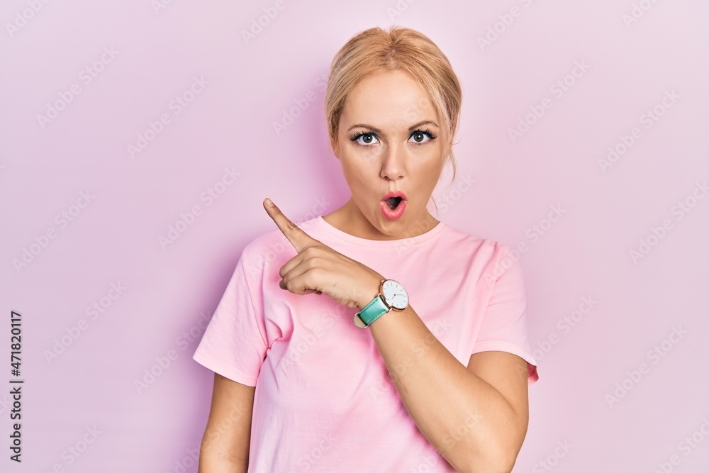 Young blonde woman wearing casual pink t shirt surprised pointing with finger to the side, open mouth amazed expression. Young blonde woman wearing casual pink t shirt surprised pointing with finger to the side, open mouth amazed expression.