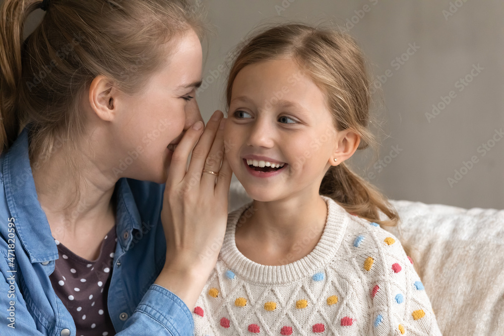 Young Caucasian mother telling secret whispering on ear of curious excited small adorable child daughter, gossiping or sharing confidential information at home, enjoying trustful conversation. Young Caucasian mother telling secret whispering on ear of curious excited small adorable child daughter, gossiping or sharing confidential information at home, enjoying trustful conversation.