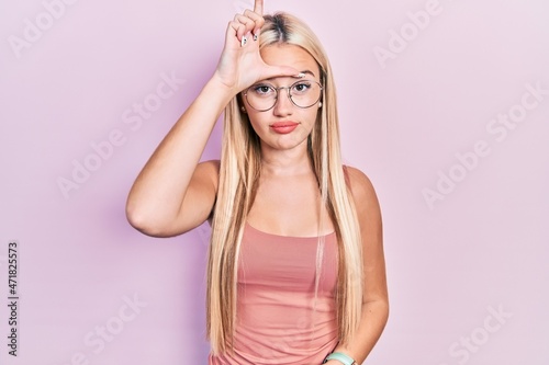 Young blonde girl wearing casual clothes making fun of people with fingers on forehead doing loser gesture mocking and insulting.