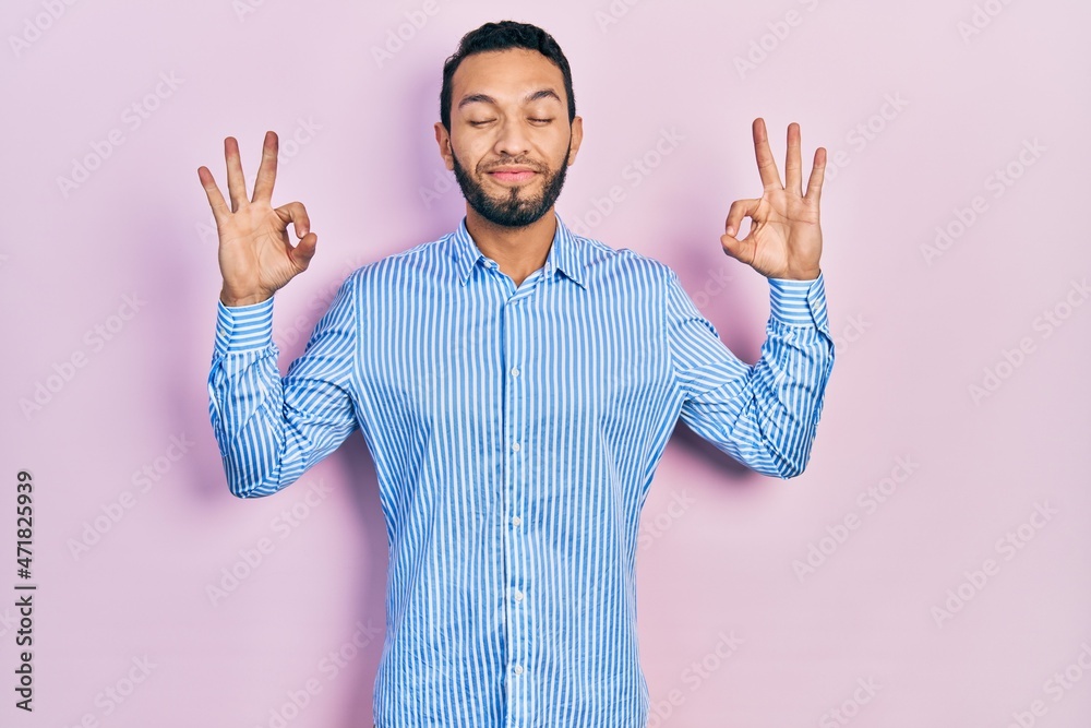 Hispanic man with beard wearing casual blue shirt relax and smiling with eyes closed doing meditation gesture with fingers. yoga concept. Hispanic man with beard wearing casual blue shirt relax and smiling with eyes closed doing meditation gesture with fingers. yoga concept.