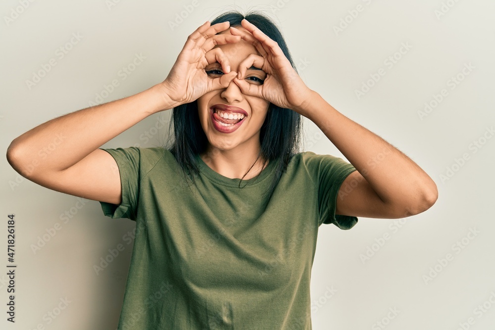 Young hispanic girl wearing casual t shirt doing ok gesture like binoculars sticking tongue out, eyes looking through fingers. crazy expression. Young hispanic girl wearing casual t shirt doing ok gesture like binoculars sticking tongue out, eyes looking through fingers. crazy expression.