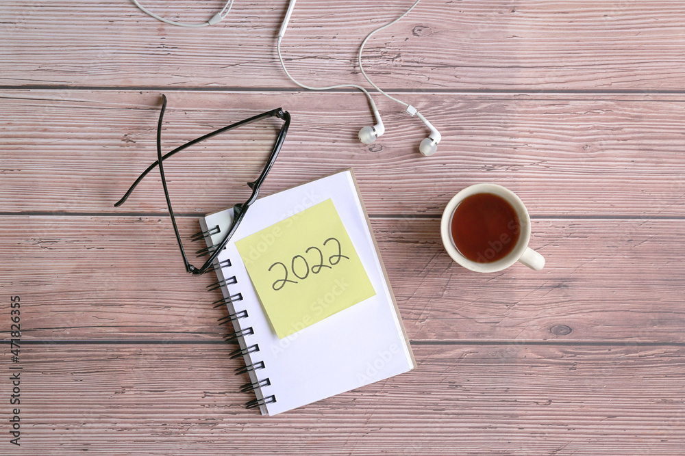 2022 on yellow paper on top of notepad with eyeglasses, coffee and ear phones on wood tabletop. Top view. 2022 on yellow paper on top of notepad with eyeglasses, coffee and ear phones on wood tabletop. Top view.