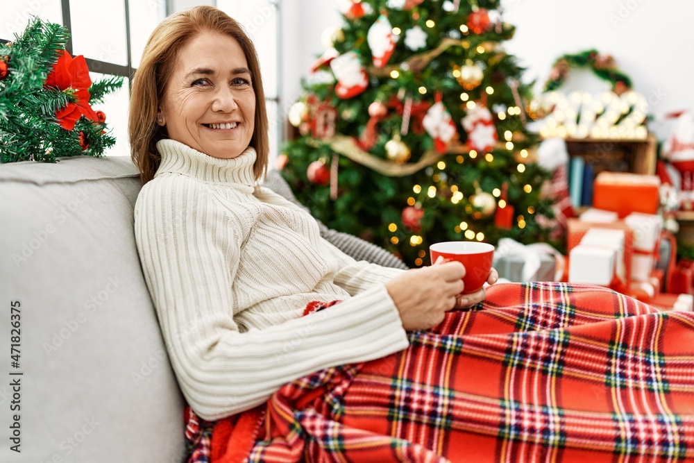 Middle age caucasian woman drinking coffee sitting by christmas tree at home