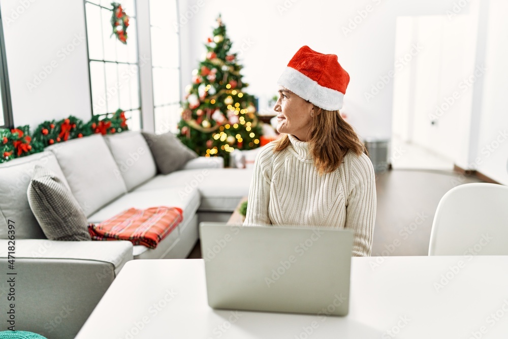 Middle age woman wearing santa claus hat using laptop looking to side, relax profile pose with natural face with confident smile. Middle age woman wearing santa claus hat using laptop looking to side, relax profile pose with natural face with confident smile.