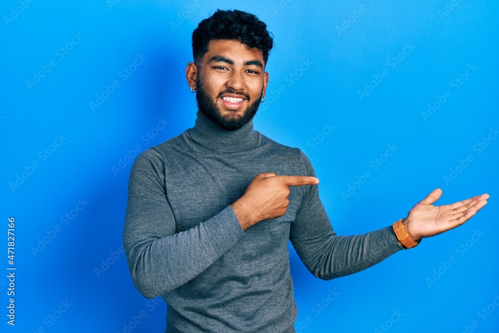Arab man with beard wearing turtleneck sweater amazed and smiling to the camera while presenting with hand and pointing with finger. Arab man with beard wearing turtleneck sweater amazed and smiling to the camera while presenting with hand and pointing with finger.