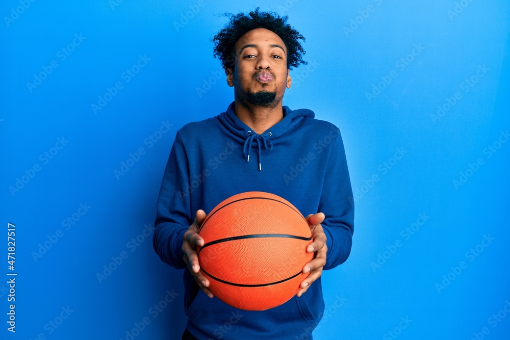 Young african american man with beard holding basketball ball puffing cheeks with funny face. mouth inflated with air, catching air.