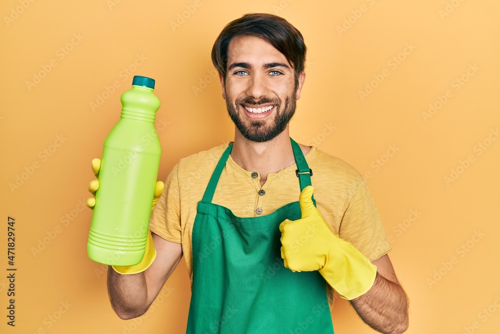 Young hispanic man wearing cleaner apron holding cleaning product smiling happy and positive, thumb up doing excellent and approval sign Young hispanic man wearing cleaner apron holding cleaning product smiling happy and positive, thumb up doing excellent and approval sign