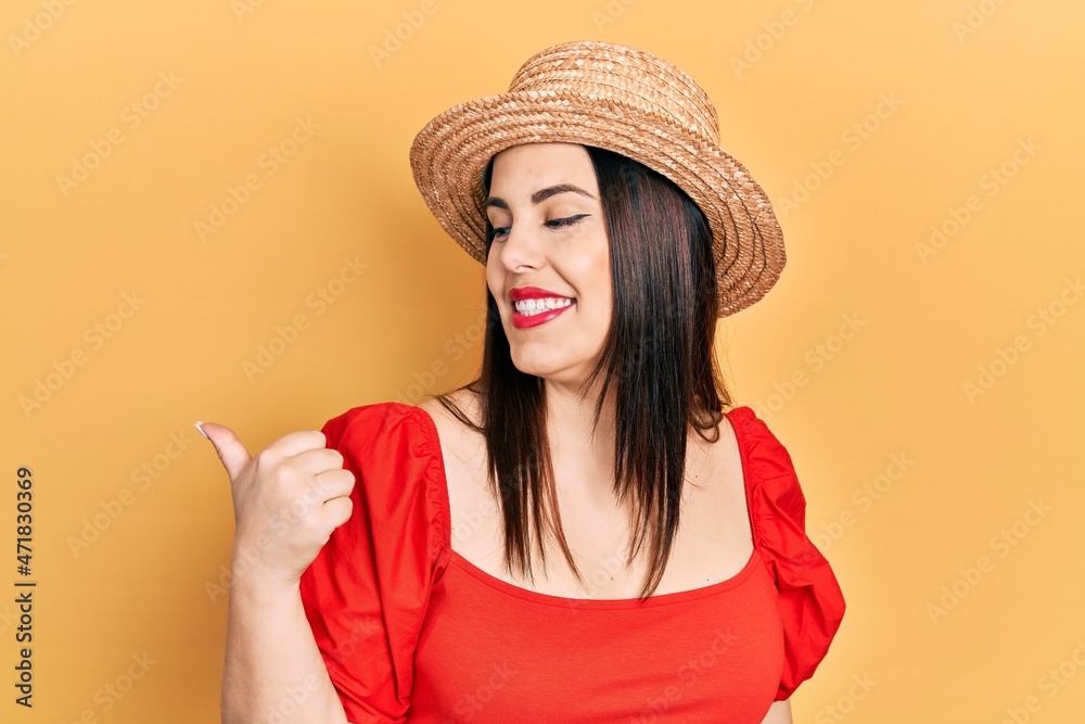 Young hispanic woman wearing summer hat smiling with happy face looking and pointing to the side with thumb up. Young hispanic woman wearing summer hat smiling with happy face looking and pointing to the side with thumb up.