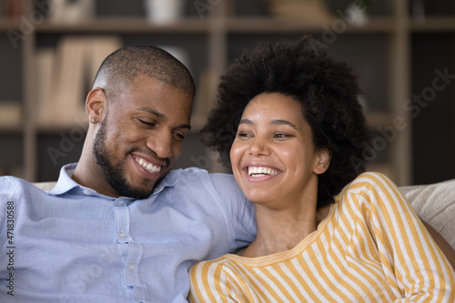 Happy African American couple relaxing at home, talking and laughing, sitting close, hugging. Young husband and wife joking and having fun, embracing on sofa, enjoying being dogether