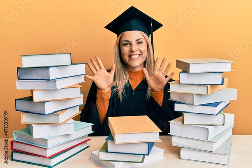 Young caucasian woman wearing graduation ceremony robe sitting on the table showing and pointing up with fingers number ten while smiling confident and happy.