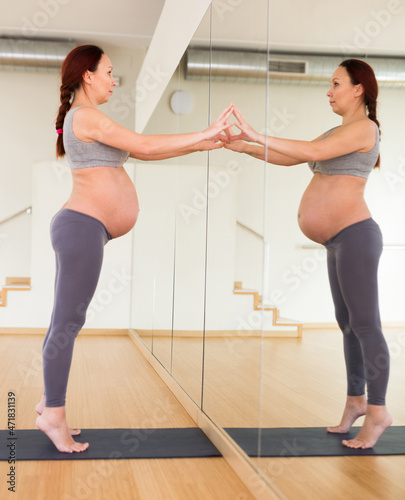 Pregnant woman doing yoga stands on tiptoe leans against the mirror wall