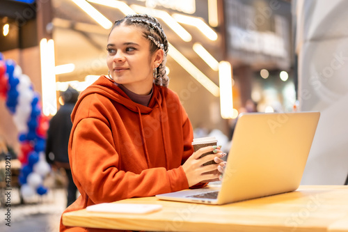 Alternative girl with white braids with a computer in a shopping center, pensive with a hot coffee in her hands