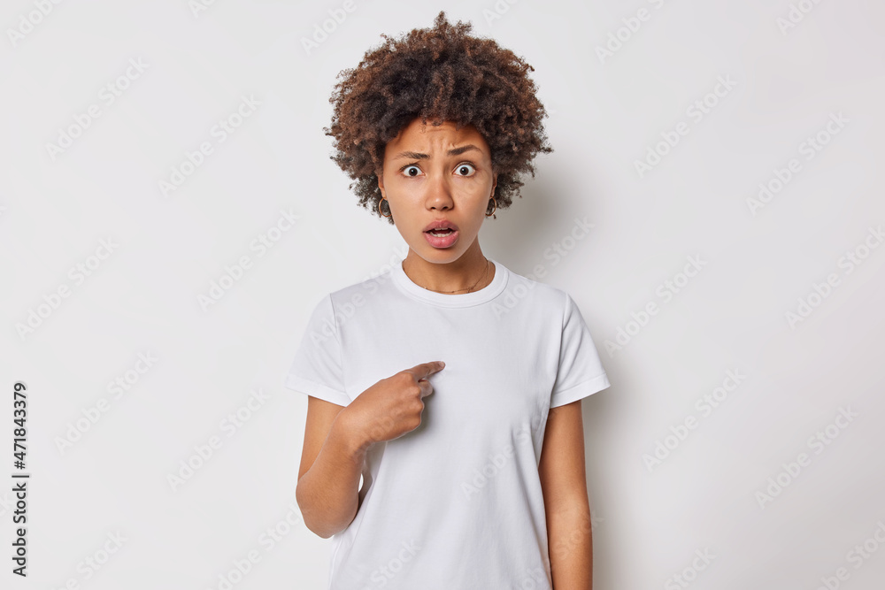 Who me. Shocked embarrassed curly haired young woman points at herself being chosen wears casual t shirt isolated over white studio background being insulted by stranger questioned being attacked. Who me. Shocked embarrassed curly haired young woman points at herself being chosen wears casual t shirt isolated over white studio background being insulted by stranger questioned being attacked.