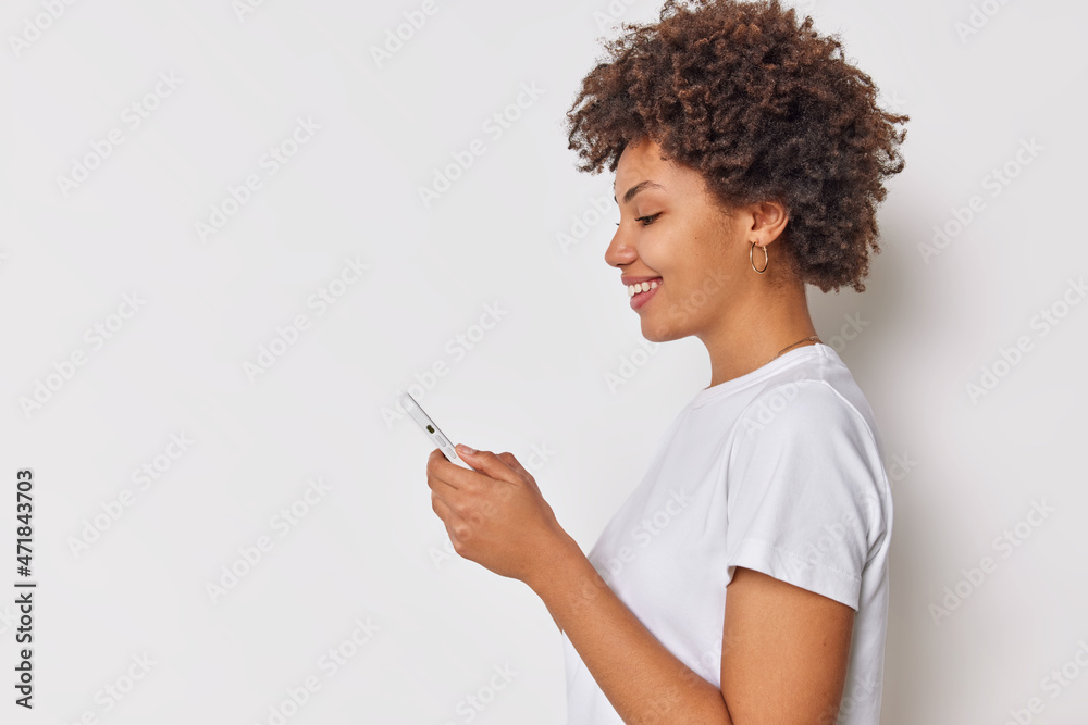 Profile shot of positive curly haired young woman uses mobile phone for communication messaging with important person dressed in casual t shirt isolated over white background blank copy space Profile shot of positive curly haired young woman uses mobile phone for communication messaging with important person dressed in casual t shirt isolated over white background blank copy space
