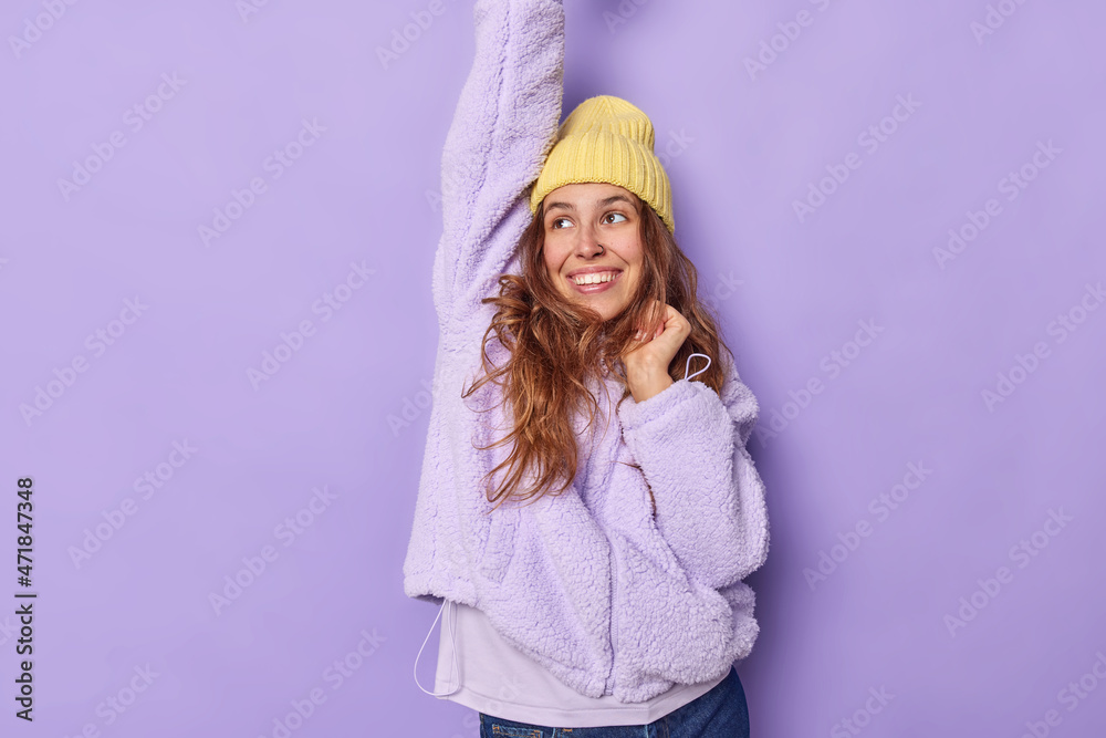 Indoor shot of happy young European woman makes triumph dance keeps arm raised dressed in warm fur jacket and yellow hat being full of joy isolated over purple background glad to achieve goal. Indoor shot of happy young European woman makes triumph dance keeps arm raised dressed in warm fur jacket and yellow hat being full of joy isolated over purple background glad to achieve goal.