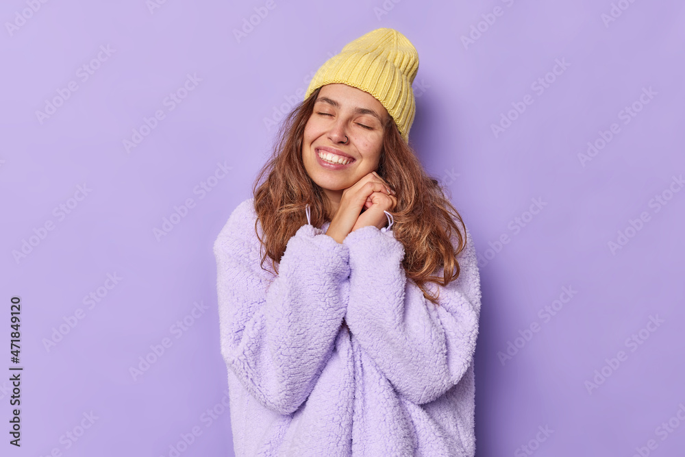 Photo of sincere young woman keeps hands near face closes eyes from satisfaction smiles gently stands tender and romantic daydreams about something dressed in outerwear isolated over purple wall Photo of sincere young woman keeps hands near face closes eyes from satisfaction smiles gently stands tender and romantic daydreams about something dressed in outerwear isolated over purple wall