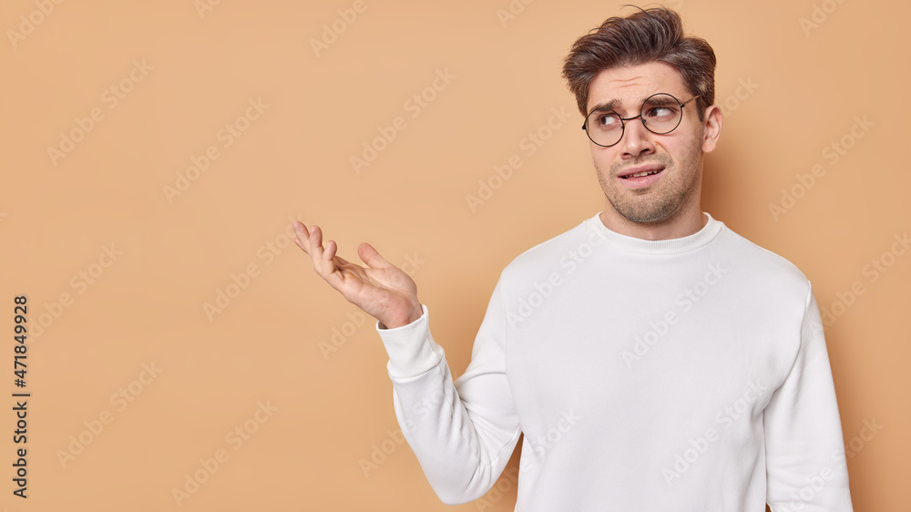 Puzzled displeased adult man has frowned expression demonstrates something umpleased keeps palm raised wears casual white jumper eyeglasses for vision correction isolated over beige studio wall Puzzled displeased adult man has frowned expression demonstrates something umpleased keeps palm raised wears casual white jumper eyeglasses for vision correction isolated over beige studio wall