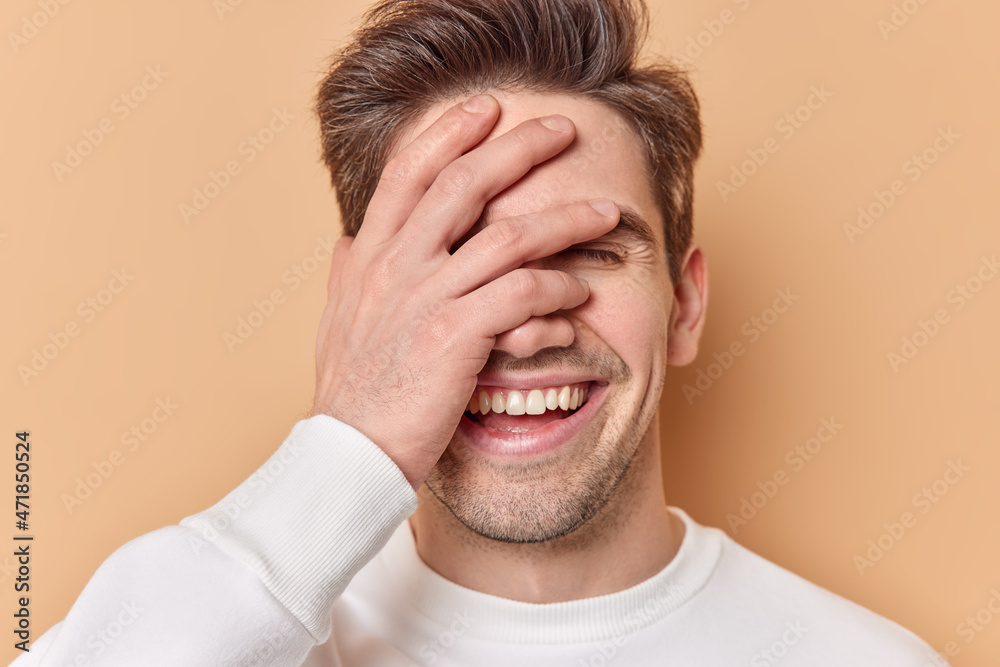 Close up shot of optimistic guy covers face with hand laughs joyfully smiles broadly hears something funny dressed in casual white jumper isolated over beige studio background hides emotions Close up shot of optimistic guy covers face with hand laughs joyfully smiles broadly hears something funny dressed in casual white jumper isolated over beige studio background hides emotions