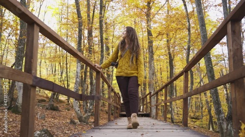 Front view woman traveller in yellow sweater with photo camera goes on wooden pathway through picturesque autumn park with colorful trees, tracking shot