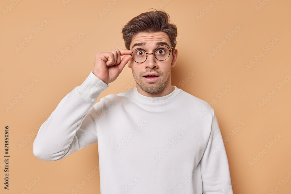 Studio shot of amazed brunet man keeps hand on rim of spectacles looks at something unbelievable cannot believe his eyes wears casual white jumper isolated over beige background. Omg concept Studio shot of amazed brunet man keeps hand on rim of spectacles looks at something unbelievable cannot believe his eyes wears casual white jumper isolated over beige background. Omg concept
