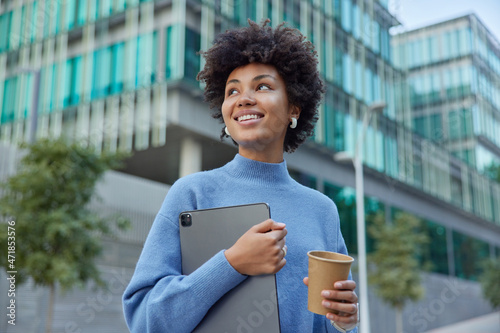Slika na platnu Positive young woman holds digital tablet uses modern gadget drinks takeaway coffee smiles happily wears casual blue jumper poses in urban setting returns from studying