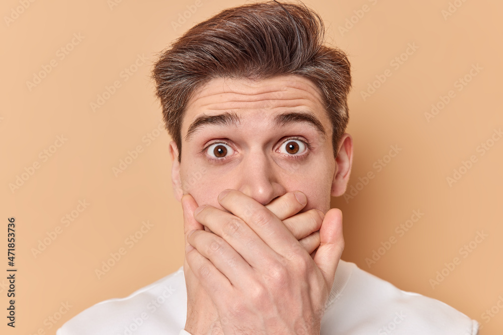 Headshot of shocked impressed young European man covers mouth with hands stands speechless tries to be speechless reacts on amazing news isolated over beige background. Human reactions concept Headshot of shocked impressed young European man covers mouth with hands stands speechless tries to be speechless reacts on amazing news isolated over beige background. Human reactions concept