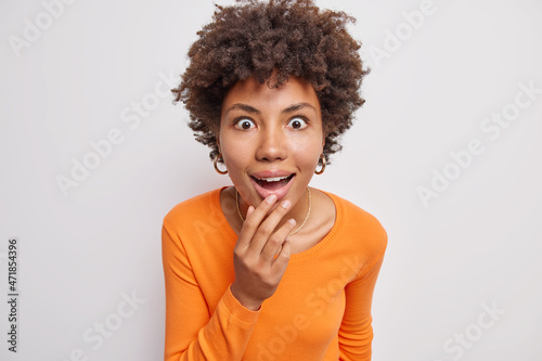 Portrait of beautiful woman hears interesting rumor gazes amazed looks intrigued keeps mouth widely opened dressed in casual orange jumper isolated over white background hears details of smth
