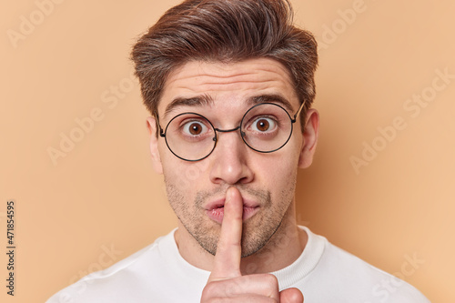 Close up portrait of mysterious handsome man keeps index finger over lips makes silence gesture gossips about something wears round spectacles isolated over beige background asks to keep secret safe photo