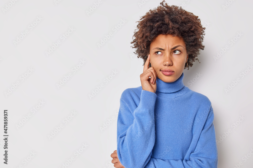 Photo of serious young curly woman keeps finger on temple tries to concentrate considers something contemplates about important thing wears casual blue jumper isolated over white background. Photo of serious young curly woman keeps finger on temple tries to concentrate considers something contemplates about important thing wears casual blue jumper isolated over white background.
