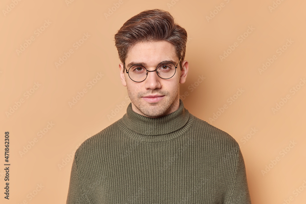 Fototapeta premium Portrait of handsome young European man looks directly at camera with serious expression feels self confident wears round transparent glasses and casual sweater isolated over beige background.
