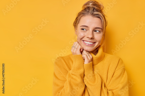 Charming pleased young woman looks tenderly aside smiles gently wears comfortable warm jumper daydreams about something isolated over vivid yellow background blank space for your advertisement photo