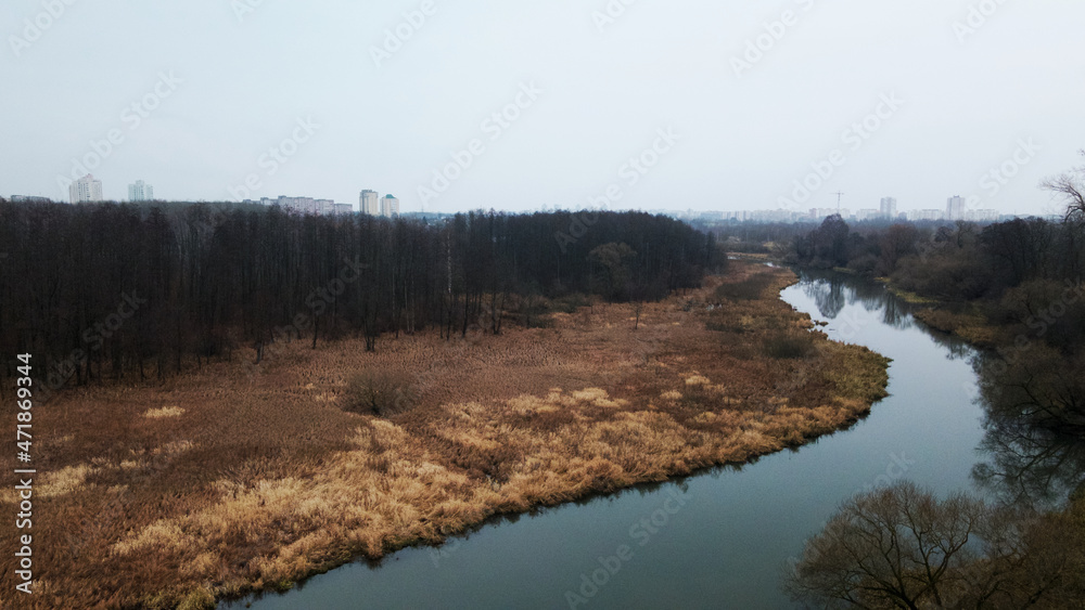 The river among the city park. Late autumn, cloudy. Trees without leaves and dried reeds along the banks of the river. The city is visible on the horizon. Aerial photography. The river among the city park. Late autumn, cloudy. Trees without leaves and dried reeds along the banks of the river. The city is visible on the horizon. Aerial photography.