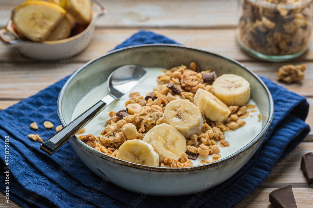 Breakfast or dessert, unsweetened yogurt with granola, dark chocolate, hazelnuts and sliced banana in a ceramic bowl on a wooden background. Breakfast recipes.