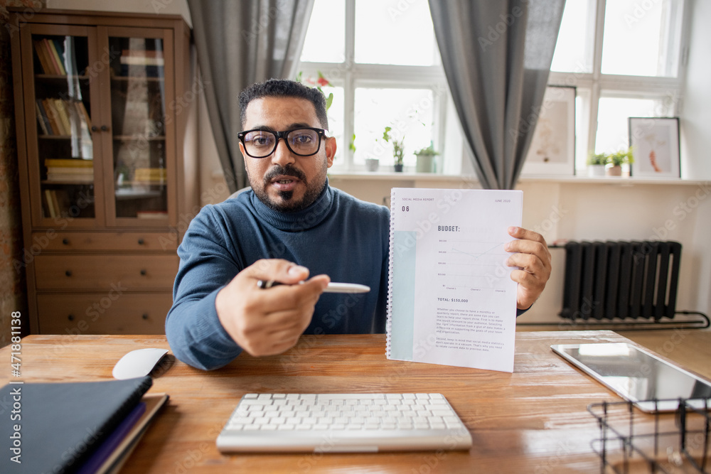 Confident teacher of economics pointing at financial document with budget graph while sitting in front of computer Confident teacher of economics pointing at financial document with budget graph while sitting in front of computer