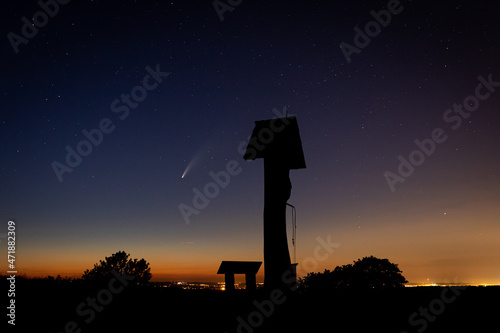 Wooden curfew bell tower on the hill with the comet Neowise, stars and the city on the background during the starlit night.