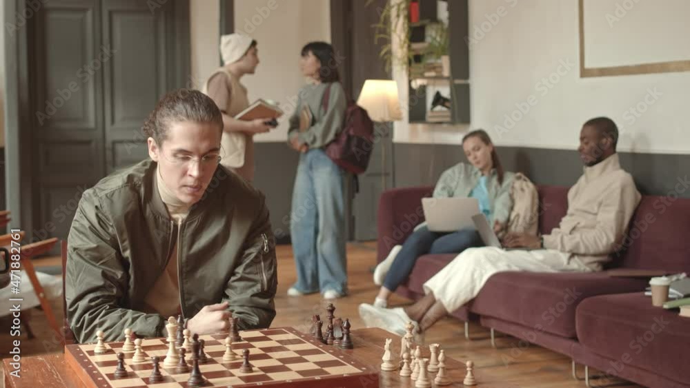 Medium slowmo shot of serious male student playing chess at modern university campus while his classmates communicating in background Medium slowmo shot of serious male student playing chess at modern university campus while his classmates communicating in background