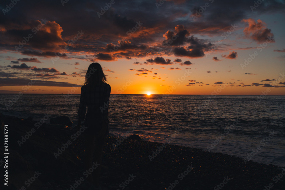 Silhouette of a young slender woman standing on the seashore and looking at a beautiful, colorful, magical sunset going beyond the horizon of the sea