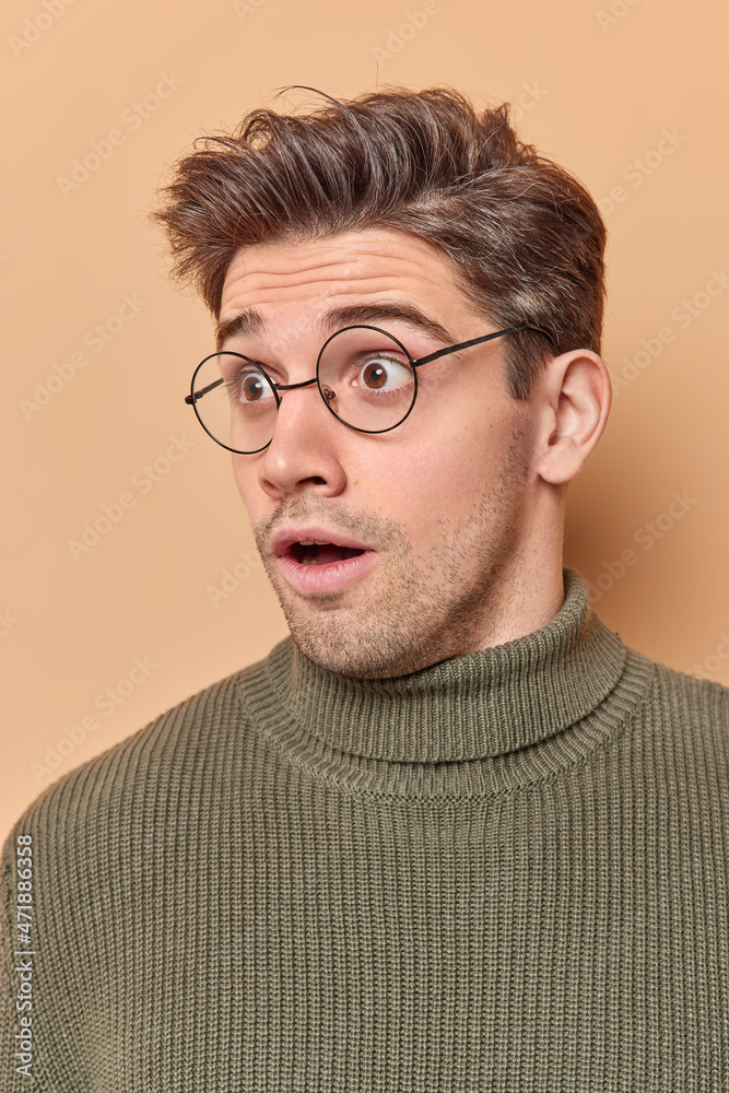 Impressed shocked adult man with dark hair stares stunned away notices horrible thing feels amazed wears spectacles and sweater reacts on amazing news isolated over beige background. Omg concept Impressed shocked adult man with dark hair stares stunned away notices horrible thing feels amazed wears spectacles and sweater reacts on amazing news isolated over beige background. Omg concept