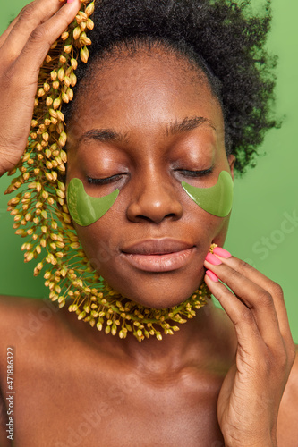 Close up shot of dark skinned young woman keeps eyes shut holds wild flower over face has healthy smooth skin stands with naked body isolated over green background. Women and beauty concept.