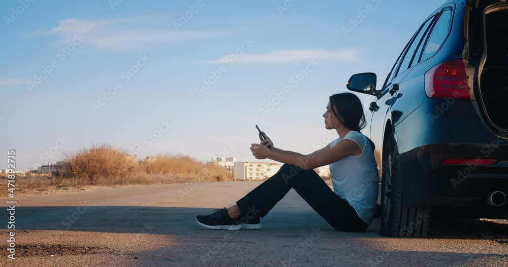 Black broken car at the emergency signal stands on the side of road with a flat tire and an upset girl. Woman answers the call from service center asking for help. Angry female can't change car tire