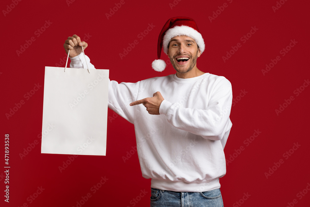 Shopping Offer. Excited Man In Santa Hat Pointing At Blank Shopper Bag Shopping Offer. Excited Man In Santa Hat Pointing At Blank Shopper Bag