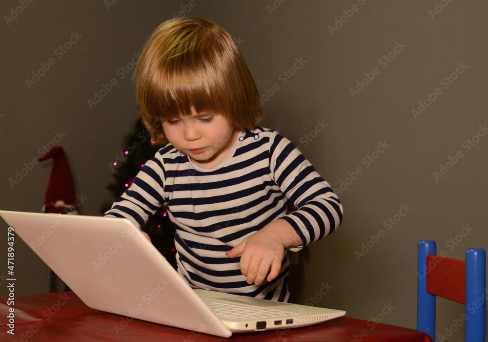 A three-year-old boy is writing a letter to Santa Claus, sitting at a laptop. against the background of the Christmas tree. Christmas concept. close-up. Gray background. A three-year-old boy is writing a letter to Santa Claus, sitting at a laptop. against the background of the Christmas tree. Christmas concept. close-up. Gray background.