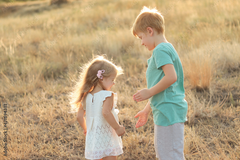 Boy and girl play together in the park. The children laugh. Brother and sister walking in a nature park among thick grass. The girl is wearing a white dress. A boy in a green T-shirt and gray shorts.