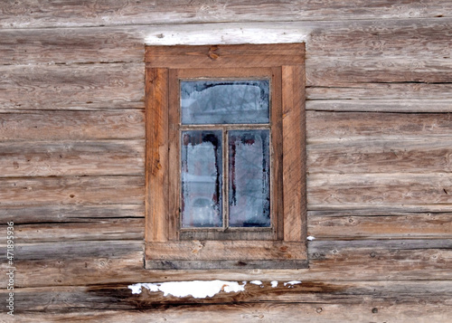 A window in a wooden frame is installed in the wall of a log cabin. Frosty patterns formed on the window glass. There is some snow on the log of the log house under the window.