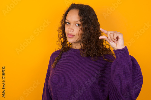 Shocked teenager girl wearing purple knitted sweater against yellow background shows something little with hands, demonstrates size, opens mouth from surprise. Measurement concept.