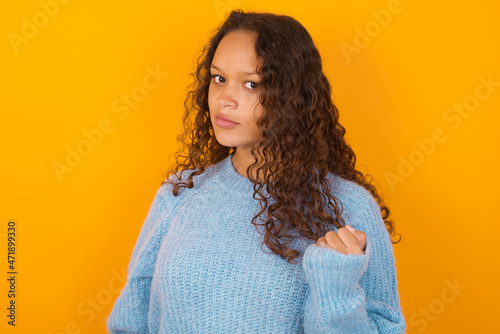 Teenager girl wearing blue sweater over yellow background shows fist has annoyed face expression going to revenge or threaten someone makes serious look. I will show you who is boss © Jihan