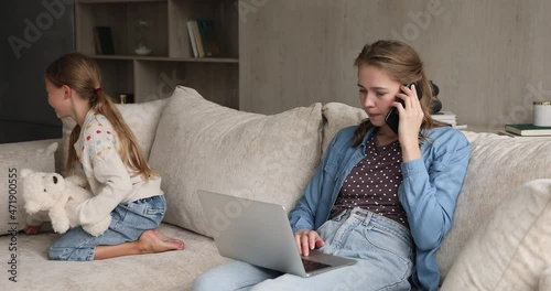 Caring young freelance mom engaged in multitasking discuss affairs on phone before pc tickle cute preteen daughter playing with toy on sofa. Businesswoman combine upbringing child and doing business