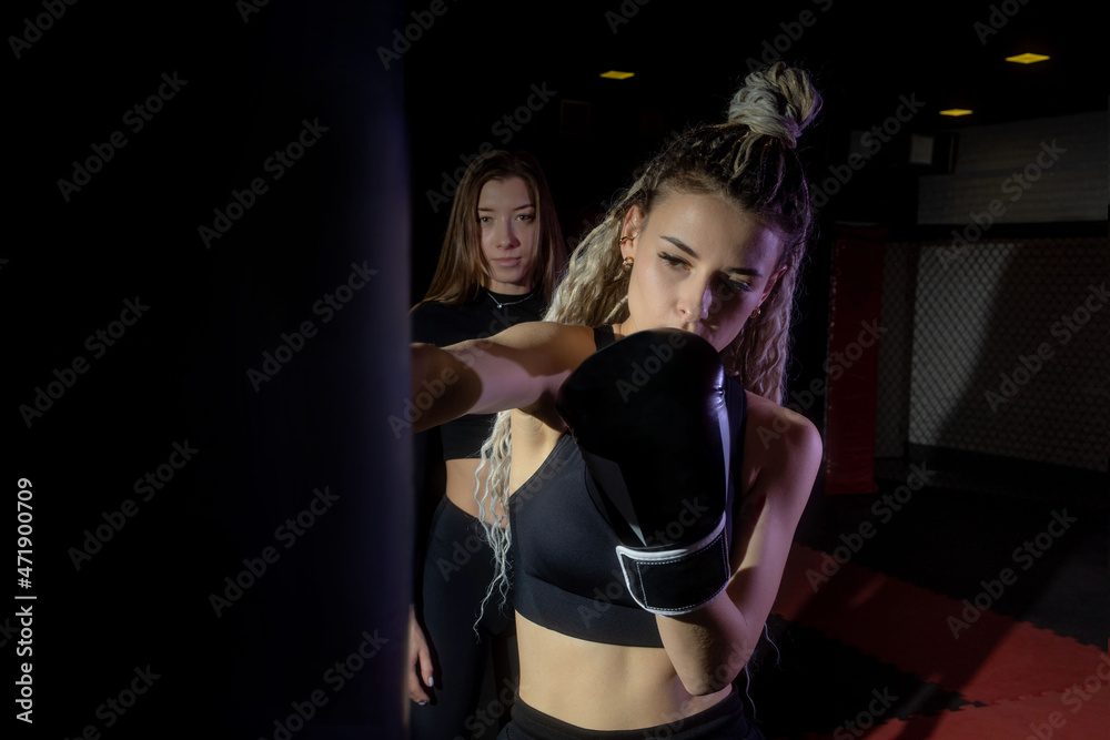A beautiful slender woman with a creative hairstyle performs the exercise - striking a punching bag while her friend watches it. A beautiful slender woman with a creative hairstyle performs the exercise - striking a punching bag while her friend watches it.