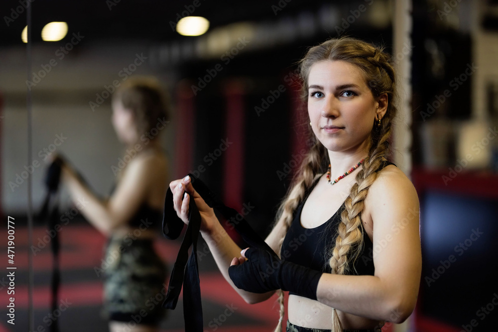 A young woman with braids prepares for a workout near big mirror, she wraps a tight bandage around her hands to protect them